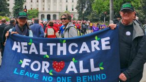 Three people hold a blue banner reading “Healthcare For All, Leave No One Behind” at a public rally. They are wearing hats and jackets, with a crowd and a large building in the background.