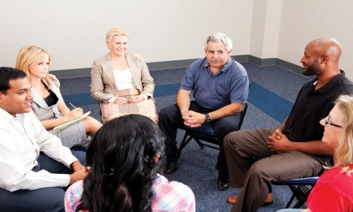 A diverse group of people seated in a circle, engaged in a discussion. The room has a neutral tone, and participants appear attentive. Some are taking notes, while others are speaking or listening.